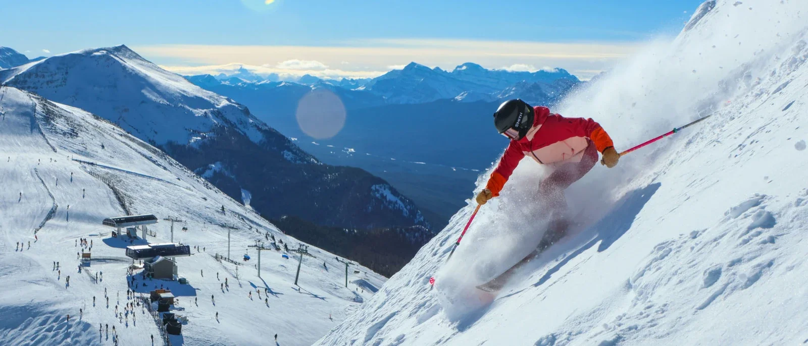 Esquiador con chaqueta roja descendiendo por nieve polvo en una pista de Banff, con remontes y montañas de las Rocosas canadienses al fondo